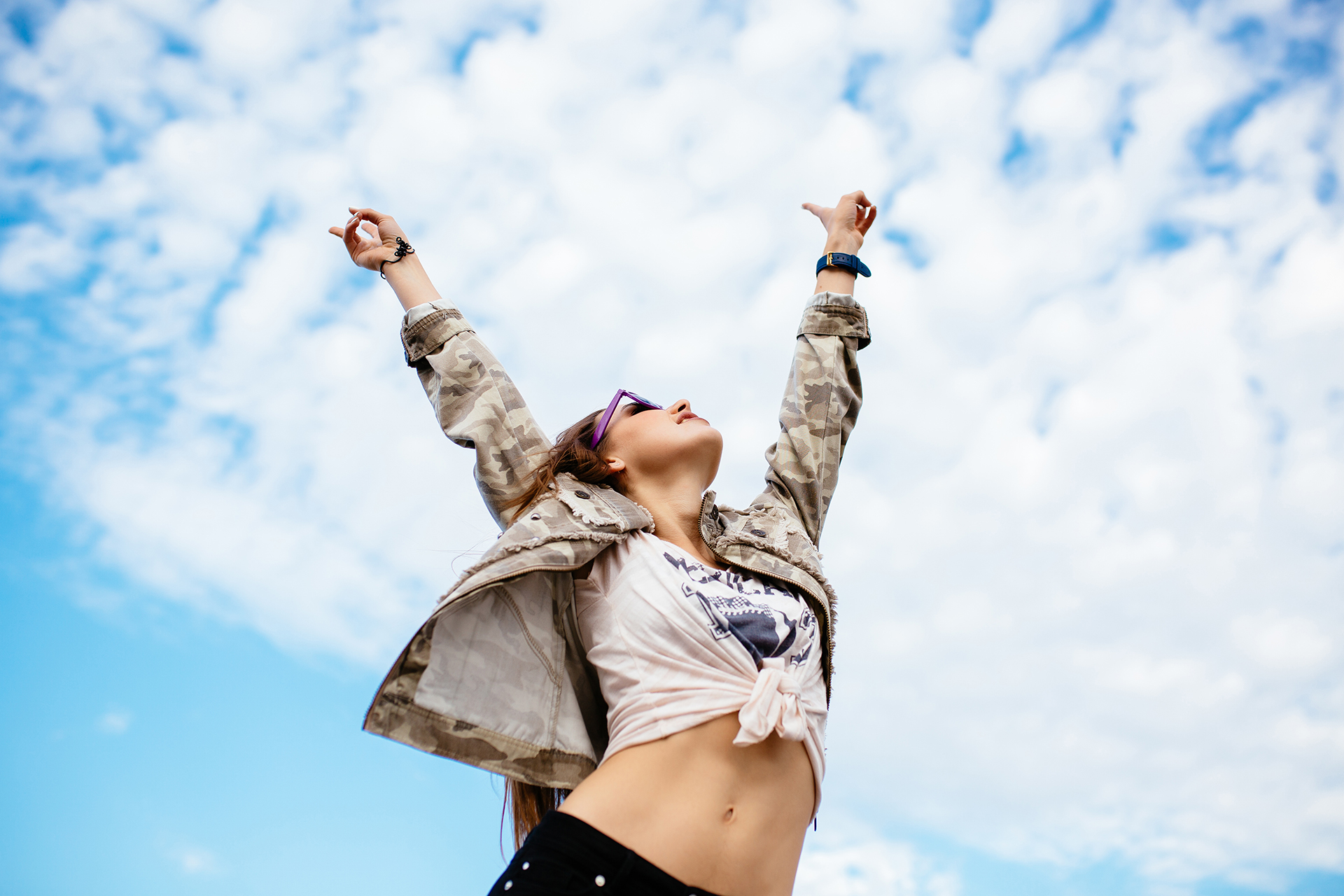 Attractive woman raised her hands up, enjoying perfect day, freedom, standing against the clear sky background, outdoors. Dressed in stylish clothes.