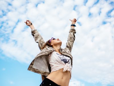 Attractive woman raised her hands up, enjoying perfect day, freedom, standing against the clear sky background, outdoors. Dressed in stylish clothes.
